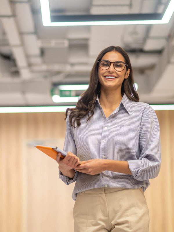 Good mood. Young long haired woman in glasses holding folder smiling at camera in illuminated room